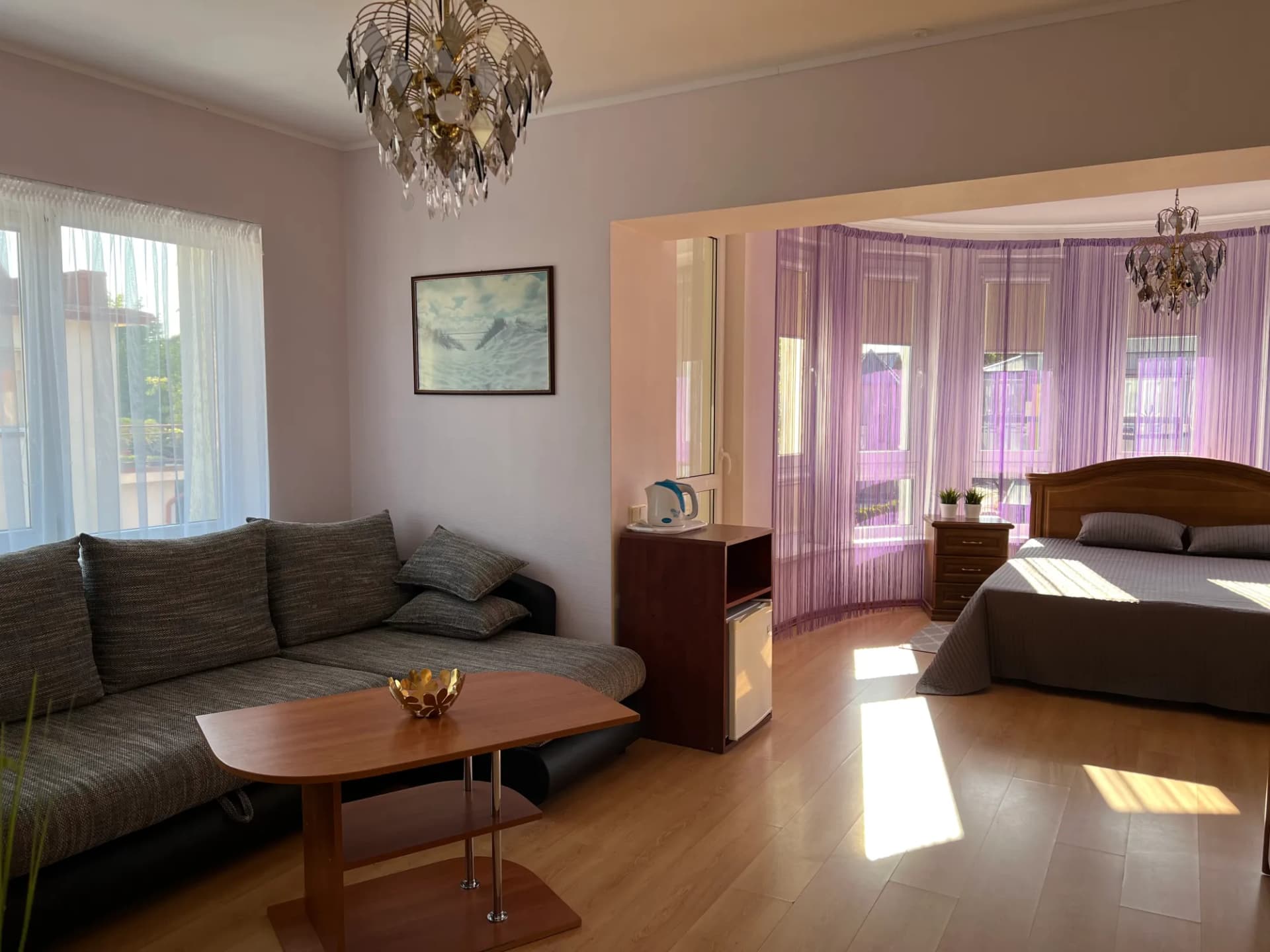 Pirmojo kambario vaizdas nuo durų: sofa, kavos staliukas ir lova prie erkerio langų