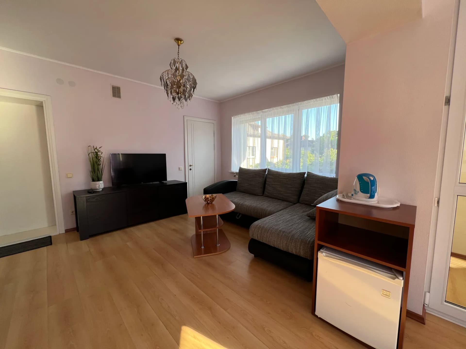 Pirmojo kambario poilsio kampas su kampine sofa, televizoriumi ir šaldytuvu